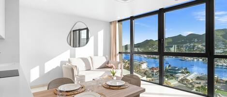A modern dining area with a wooden table set for four, large windows offering a view of a marina and hills.