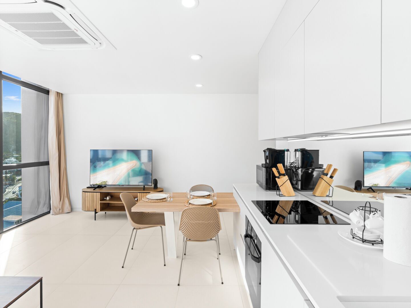 A modern kitchen with two TVs, a dining area with a wooden table and chairs, and a large window with a city view.