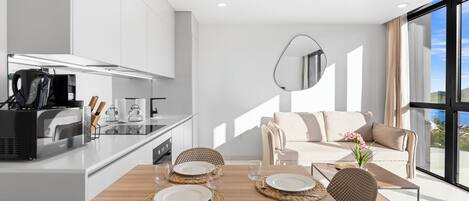 Uma cozinha moderna com fogão embutido, um espaço de refeições com mesa de madeira e jogos americanos trançados, e um sofá com vista para o exterior.
