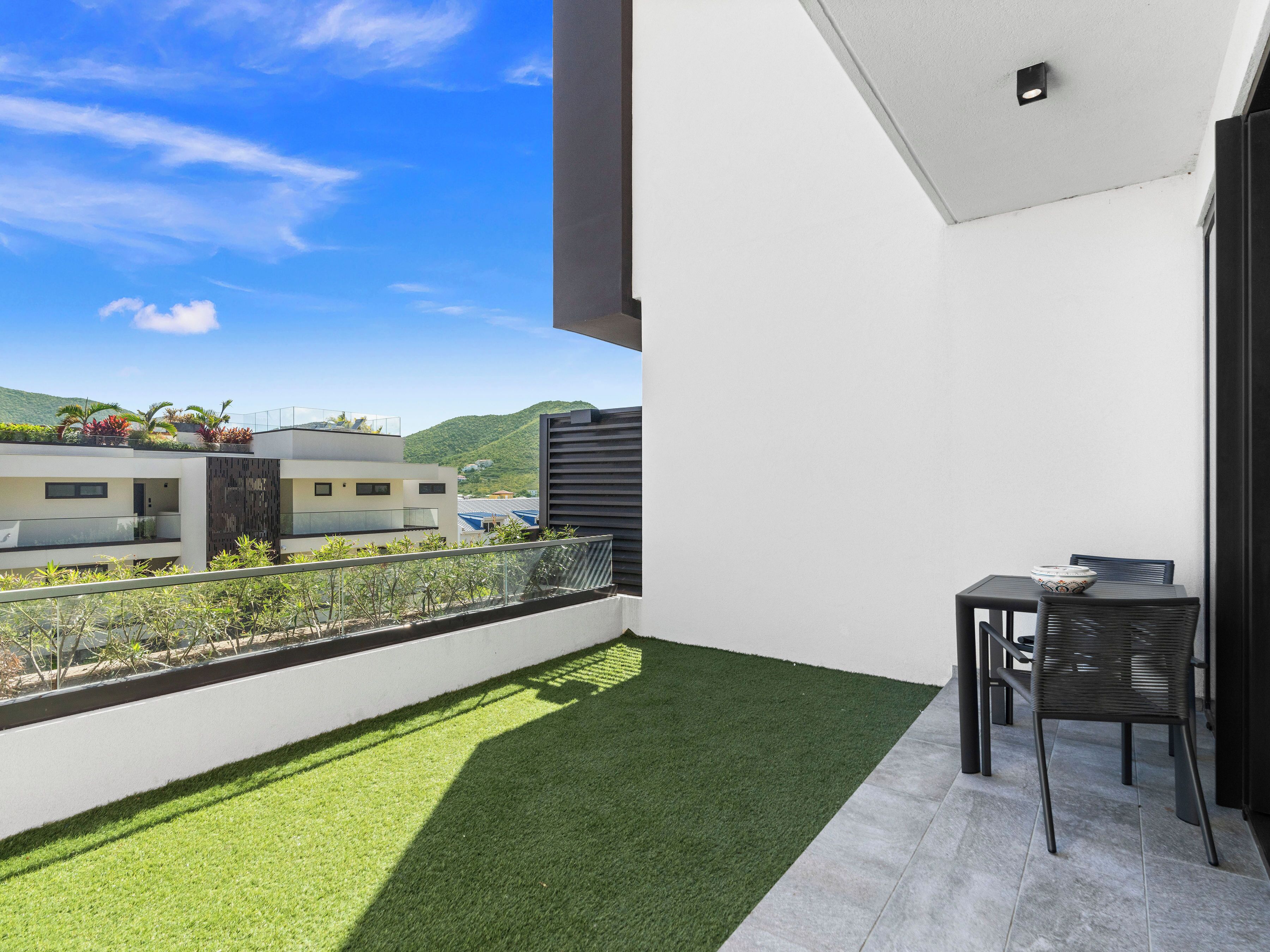 A balcony with artificial grass, a table with a chair, and a view of neighboring buildings and a hill.