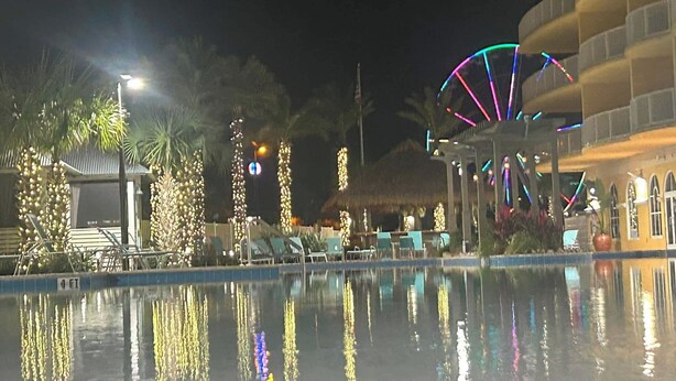 Vista nocturna de un área de piscina de hotel, mostrando el reflejo de luces en el agua, sillas de descanso y un edificio con techo curvo.