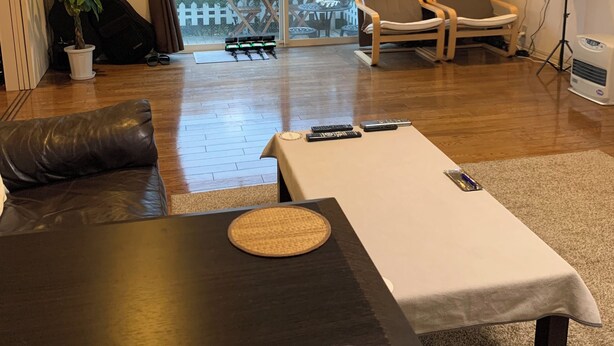 A hotel room with a wooden floor, a sofa, a table with a coaster, a chair, a balcony with a view of greenery, and an air conditioning unit. - Large 3-bedroom house in Ota-ku suburb with garden. Close to city and airport. (Ota City)
