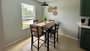 Un espace repas comprenant une table, des chaises, un luminaire suspendu, une fenêtre avec des stores et des éléments de décoration murale.