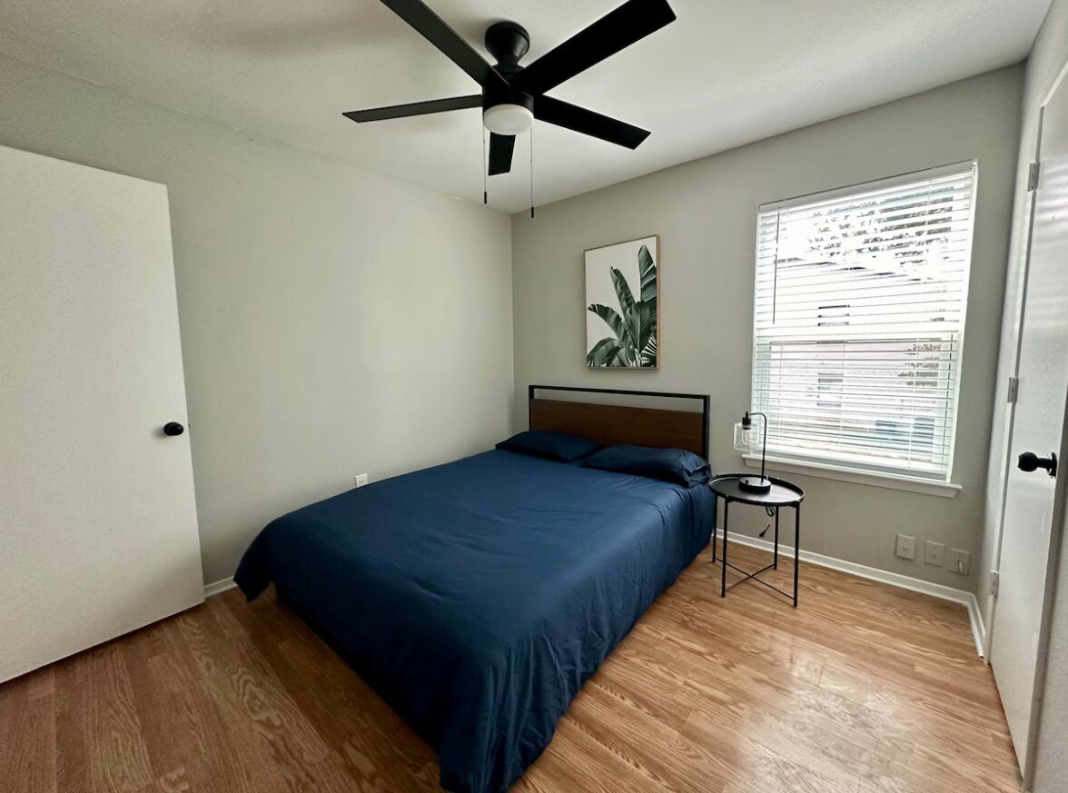 Une chambre avec un lit, un ventilateur de plafond, une fenêtre avec des stores et une petite table.