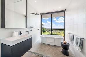 A modern bathroom with a large bathtub, a separate shower area, a vanity with a sink, and a large window offering a view of a garden and landscape.