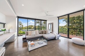 Un salon moderne avec un canapé d’angle, une table basse en verre et de grandes fenêtres donnant sur un paysage verdoyant.