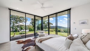 Een slaapkamer met een groot bed, een plafondventilator en uitzicht op een tuin en heuvels.