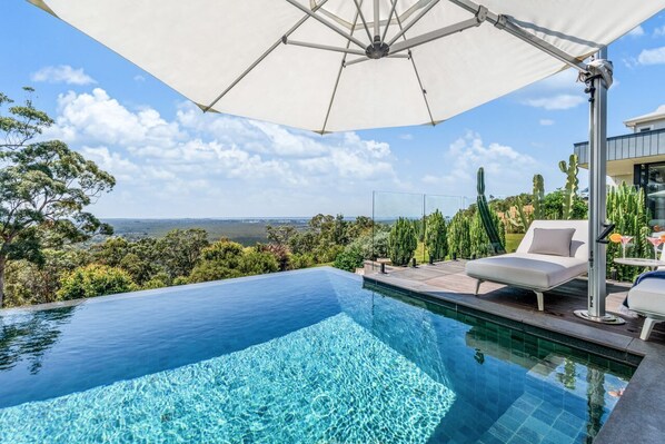 Une piscine à débordement offrant une vue imprenable sur le paysage, un parasol blanc et un fauteuil de relaxation.