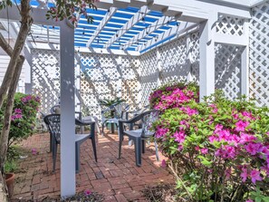 A patio area with a white lattice structure, a table with chairs, and vibrant pink flowering plants.