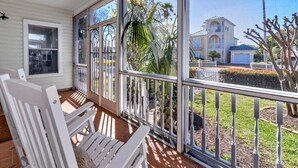 A covered porch with white chairs, large windows, and a view of a residential area with palm trees and a house. - Charming Destin Home | Yard, Pool & Beach Access (Destin)
