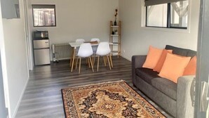 A modern living room with a grey sofa, orange cushions, a patterned rug, a dining area with white chairs, and a kitchen with a fridge and radiator.