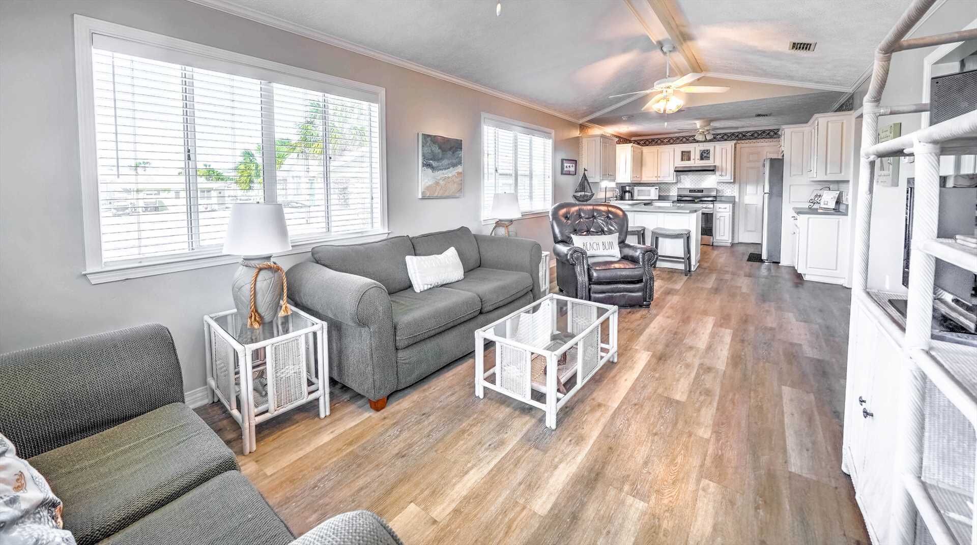 A spacious living room with a grey sofa, a coffee table, and a leather armchair. A kitchen with white cabinets and a dining area is visible in the background.