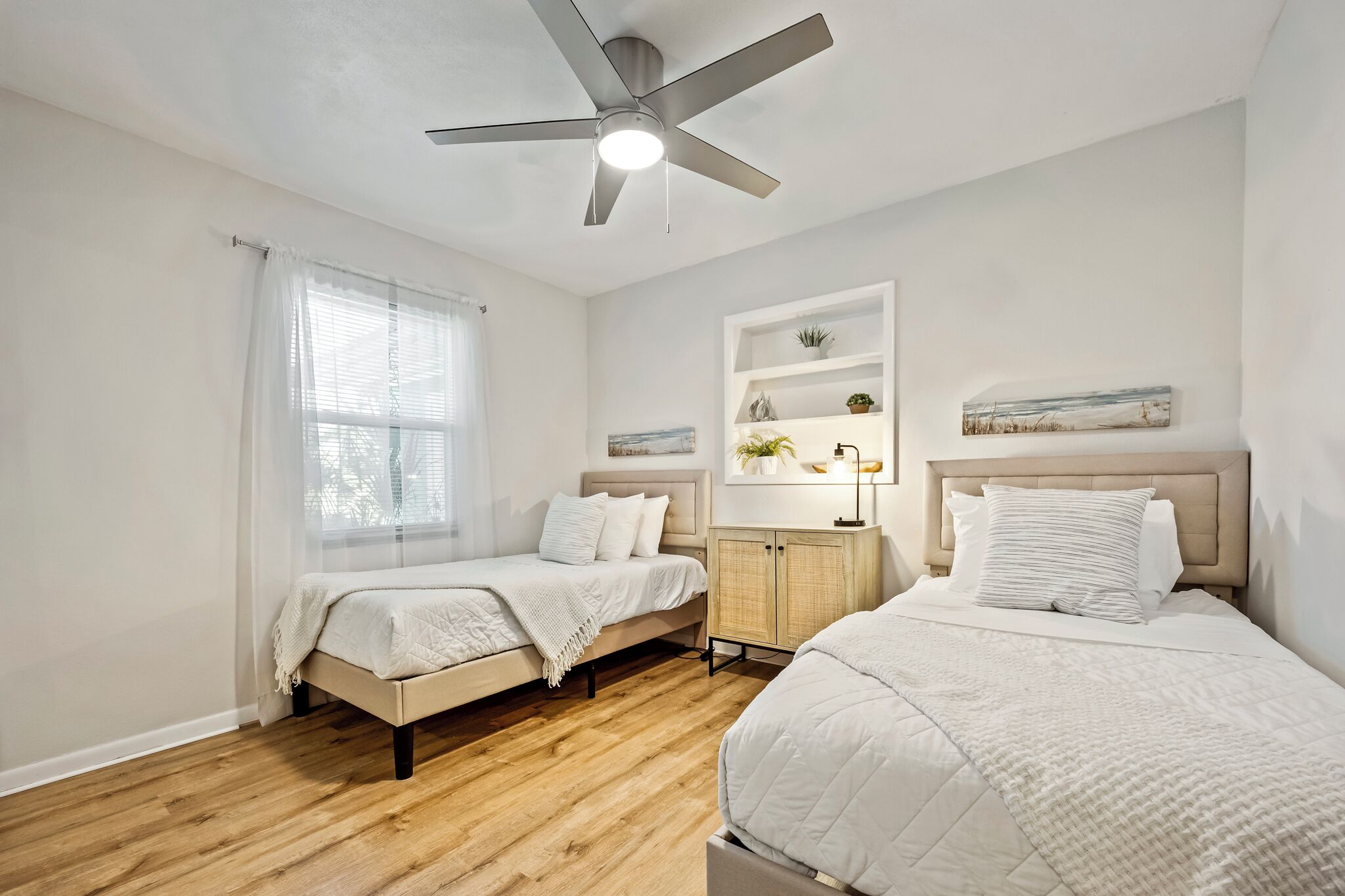 A bedroom with two beds, a ceiling fan, a window with sheer curtains, a bedside table with a lamp, and a wall-mounted shelf with decorative items.