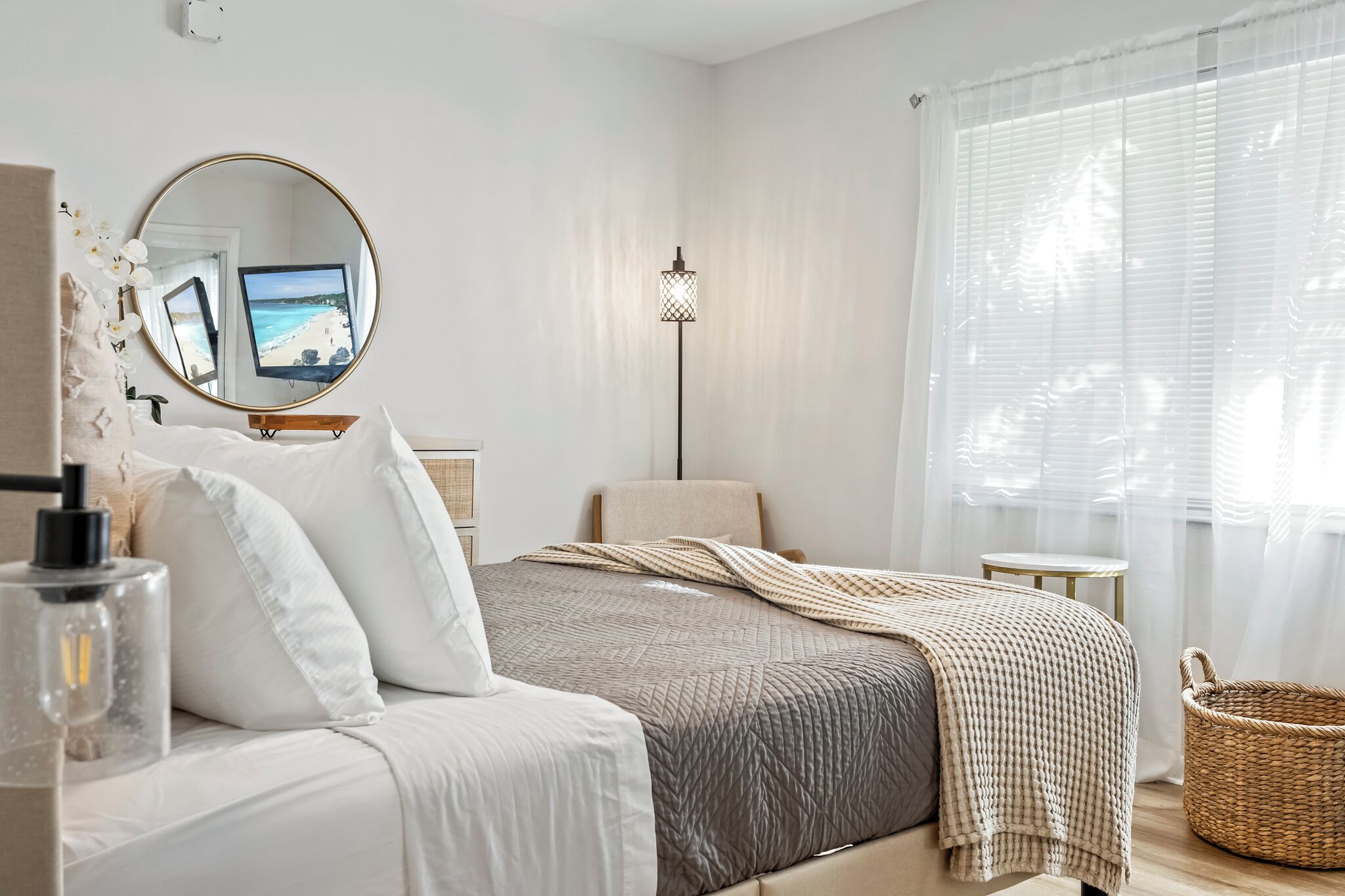 A bedroom with a bed, a bedside table, a lamp, a mirror, and a window with sheer curtains.