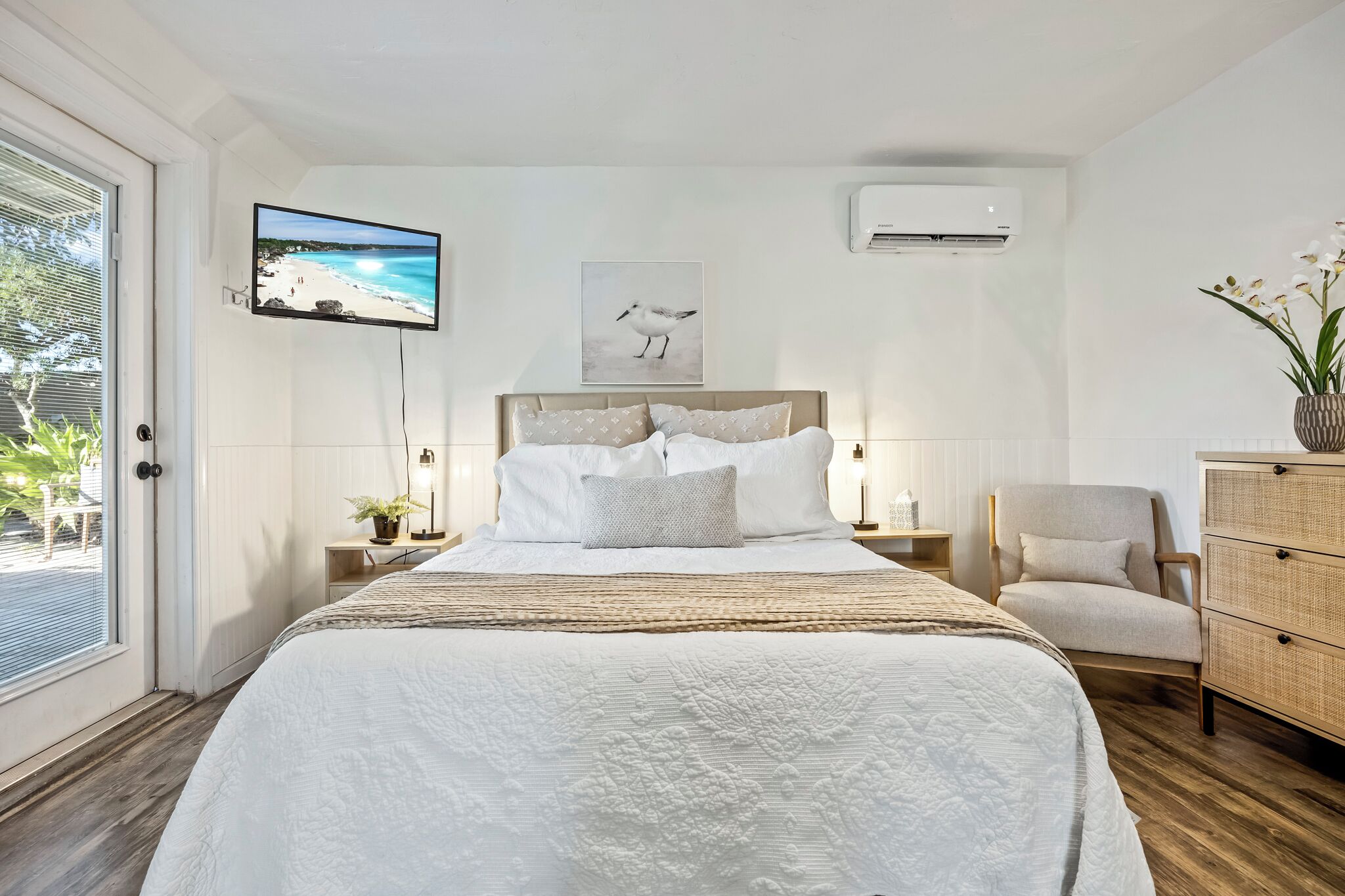 A bedroom with a large bed, a television mounted on the wall, an air conditioning unit, a chair, and a dresser.