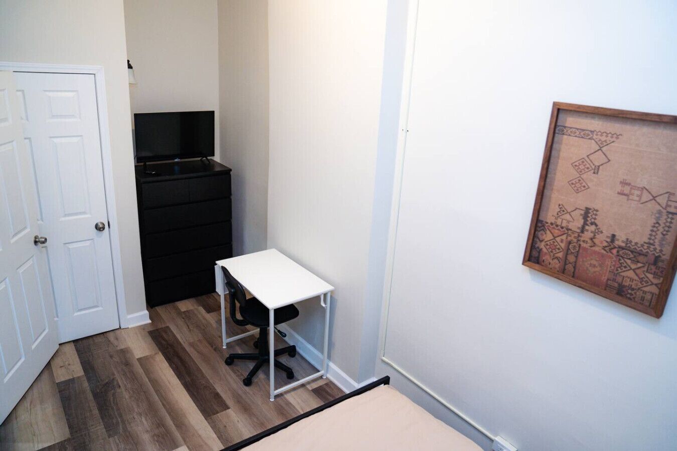 A small room with a white desk, a black chair, a dresser, and a framed picture on the wall.