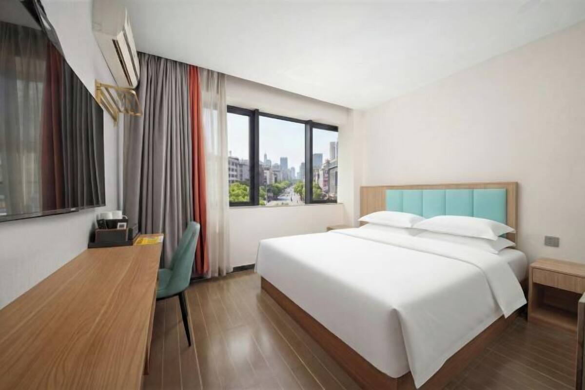 Habitación de hotel con cama, escritorio, silla, mesita de noche, ventana con vistas a la ciudad y cortina.
