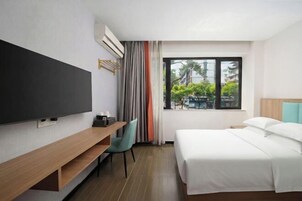 Habitación de hotel con cama, televisor, escritorio y ventana con vista a edificios y árboles.