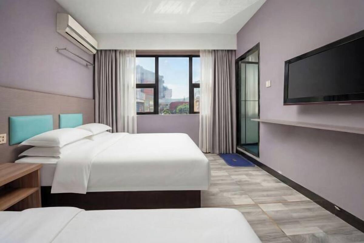 Habitación de hotel con cama, televisor, ventana con cortinas y vista a edificios.