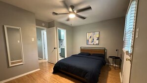 A bedroom with a bed, a ceiling fan, a painting, and a door leading to another room.