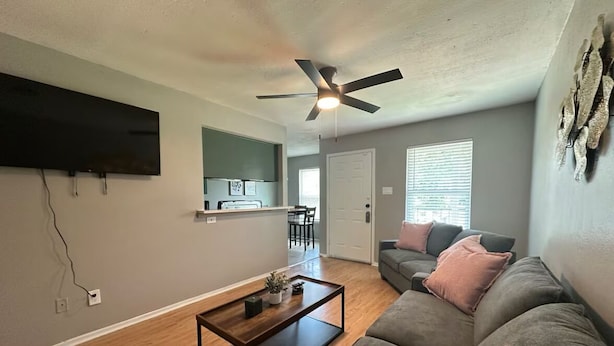 A living room with a flat-screen TV, a ceiling fan, a wooden coffee table, a gray sofa, and a door leading to a kitchen.