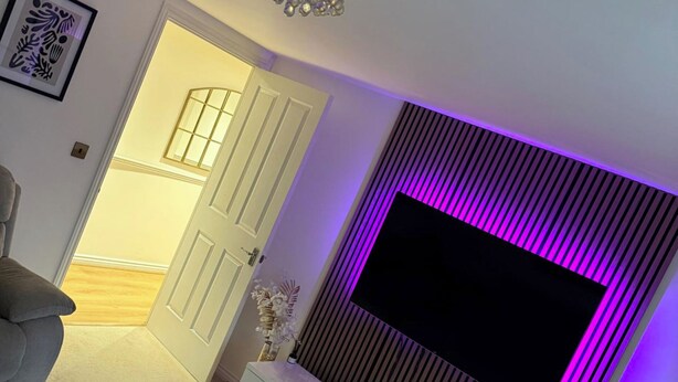 A modern living room with a flat-screen TV mounted on a wall with vertical slats, a white door, and a grey sofa. - Stylish Apartment in Chafford Hundred (Grays)