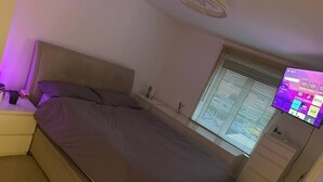 A bedroom with a bed, a nightstand, a dresser, a window with blinds, and a mounted TV displaying a home screen. - Stylish Apartment in Chafford Hundred (Grays)