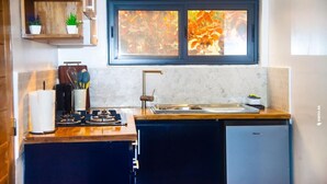 A compact kitchen with a built-in stove, a sink, and a compact refrigerator. There is a window above the sink with a view of autumn leaves. - Loft VIIM | Les Ferys du lac (Ouidah)