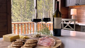Una tabla de madera con queso, galletas y jamón sobre una encimera de cocina. Una botella de vino está en la encimera, y una ventana con vistas a árboles se ve al fondo.