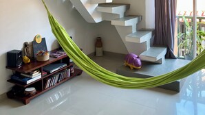 Una sala de estar moderna con una escalera blanca, una hamaca verde, una estantería y un amplio ventanal con cortinas.
