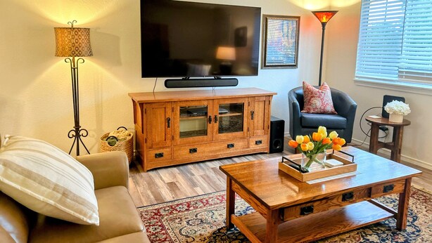 Een woonkamer met een flatscreen televisie, een houten kast, een bank, een salontafel met bloemen en een kamerbreed tapijt.