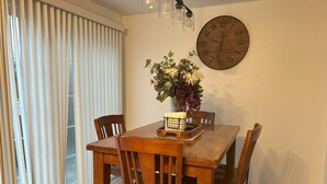 Een eetgedeelte met een houten tafel, vier stoelen, een grote wandklok en een vaas met bloemen.