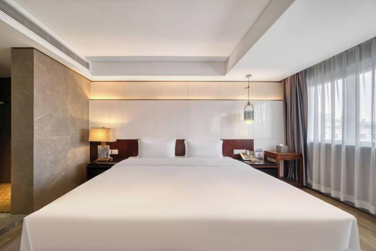 Ein Hotelzimmer mit einem großen Bett, Nachttischen, einer Lampe und Blick auf die Stadt durch das Fenster.