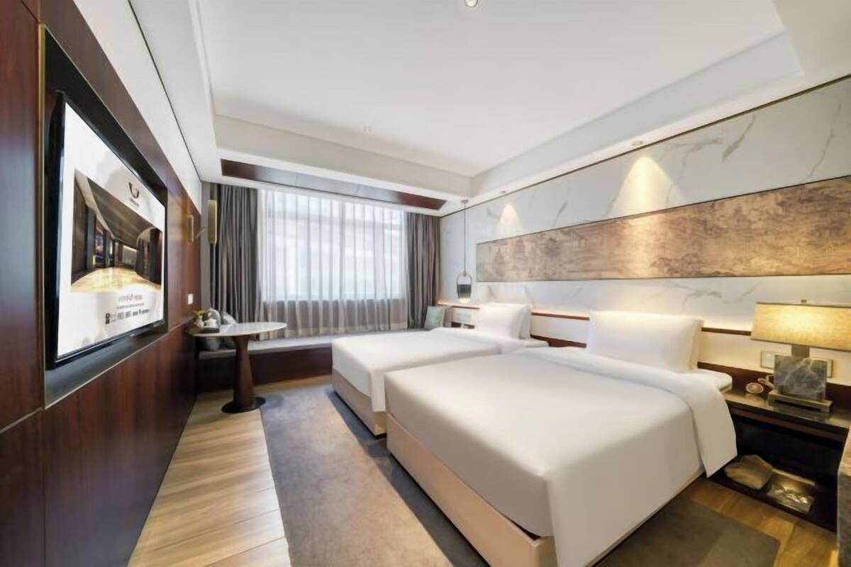 Ein Hotelzimmer mit zwei Betten, einem Schreibtisch, einem Sessel, einem großen Fenster mit Vorhängen und einem am Wand befestigten Fernseher.