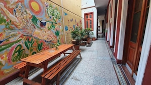Un pasillo estrecho con un banco de madera y una mesa, plantas en macetas y un mural colorido en la pared.
