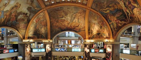 A spacious shopping mall with multiple levels, featuring a grand ceiling with intricate frescoes and a central fountain area.