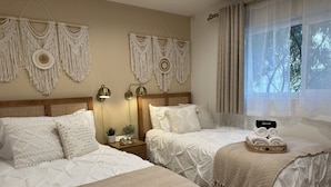 A hotel room with two beds, a bedside table with a lamp, a wall-mounted macrame decoration, and a window with curtains.