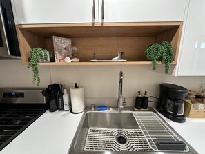 Une cuisine équipée d’un évier en acier inoxydable, d’une machine à café et d’une étagère sur laquelle se trouvent une plante et une boîte de friandises glacées.