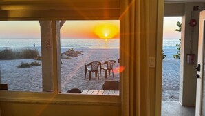 A room with a view of a beach at sunset, featuring a wooden table and chairs, a window with a view of the ocean, and a door leading outside.