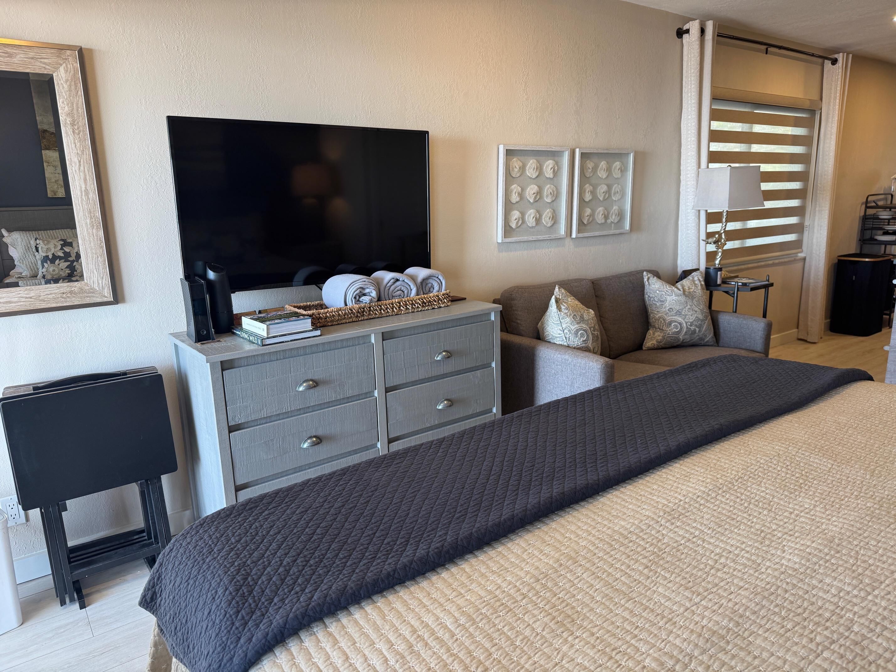 Habitació d'hotel moderna amb un llit gran, un sofà gris, una televisió de pantalla plana, un escriptori petit i una finestra amb persianes.