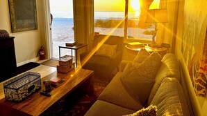 A living room with a view of the ocean at sunset, a wooden coffee table, a sofa, and a lamp.