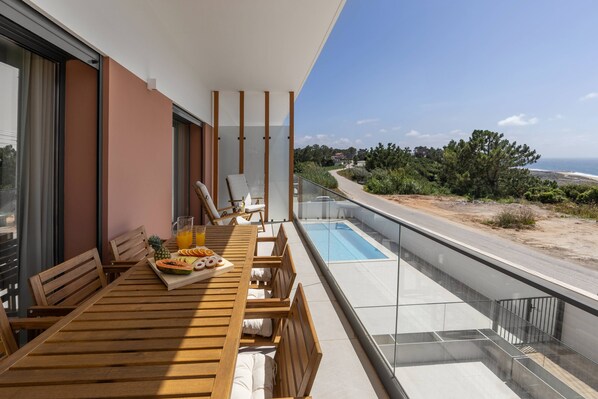 A balcony with a wooden table set for breakfast, a glass railing, and a view of the beach and ocean.