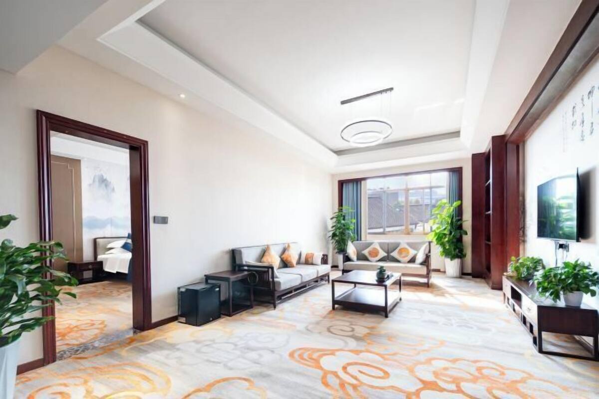 A modern living room with a sofa, coffee table, and a TV. There are plants and a large window.