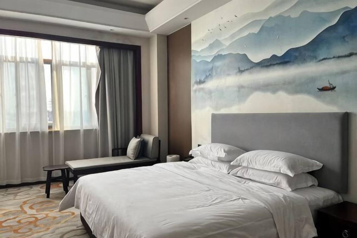 A hotel room with a large bed, a bedside table, a chair, a window with curtains, and a wall mural.