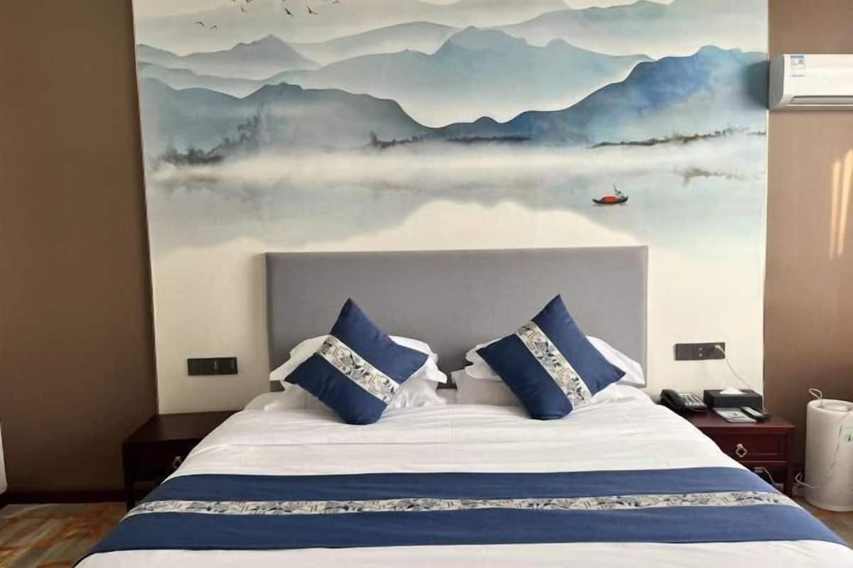 A hotel room with a bed, pillows, a nightstand, a lamp, and a wall painting.