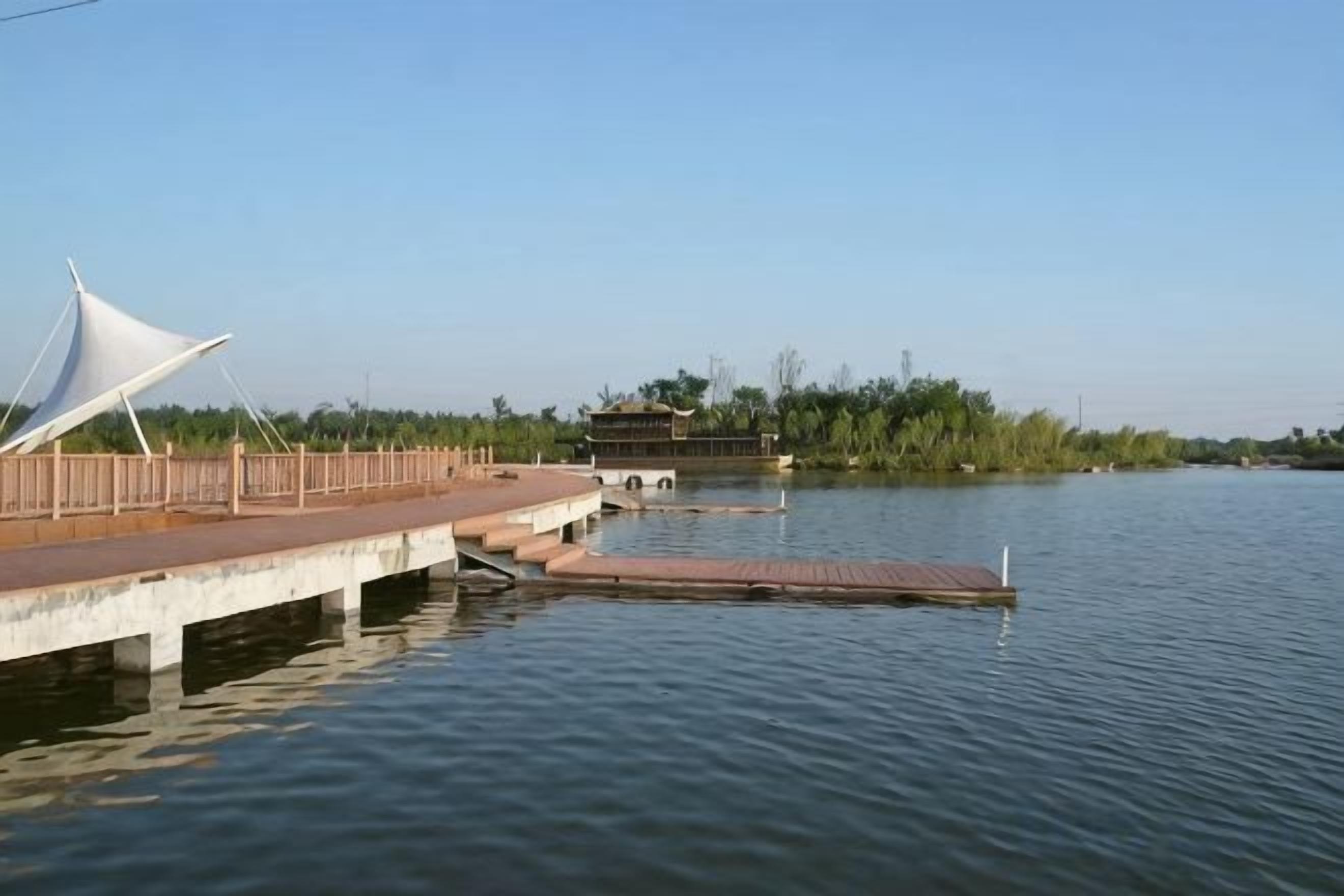 Un paseo marítimo con una estructura blanca tipo marquesina, una pasarela de madera y un edificio a lo lejos, al otro lado del agua.