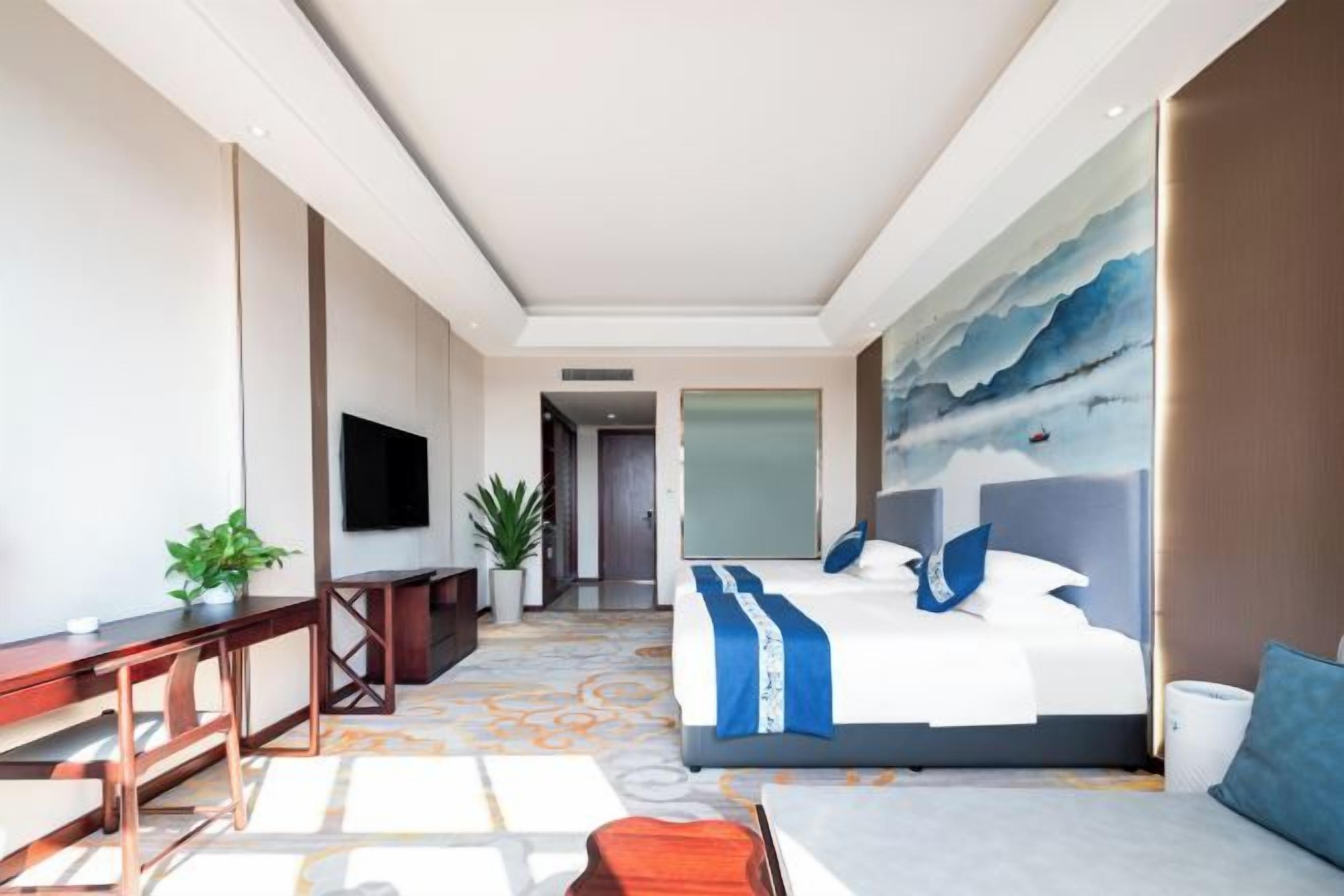 Habitación de hotel con cama, escritorio, televisión, planta y un cuadro en la pared.