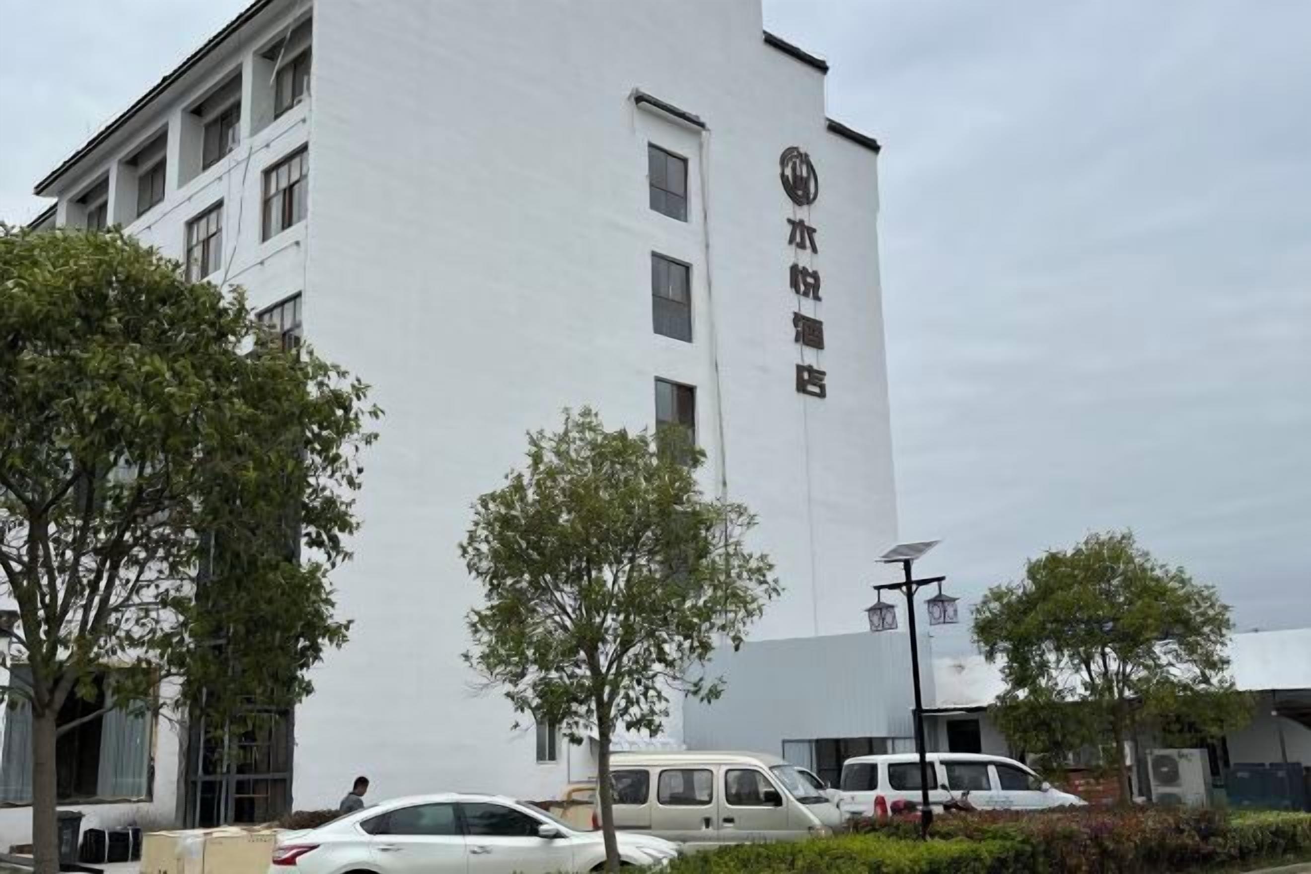 A multi-story building with a white facade, several windows, and a sign with Chinese characters. There are trees in front of the building and a few cars parked nearby.