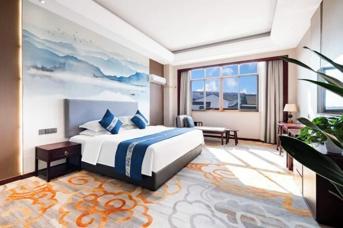 A hotel room with a bed, a desk, a chair, a large window, and a painting on the wall.