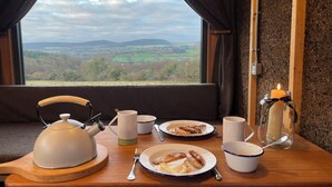 Een ontbijttafel met borden eten, een waterkoker, kopjes en een kaars, met op de achtergrond een mooi landschap.